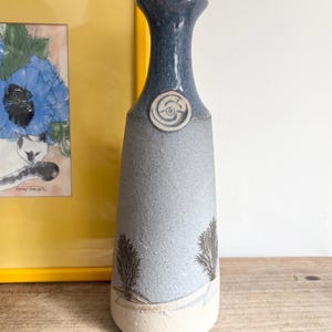 May include: A tall ceramic vase with a tapered neck and a flared rim. The upper section is a glossy dark blue, while the lower body features a matte light grey finish with two dark brown tree silhouettes and a white band at the base.