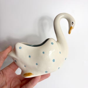 May include: A ceramic swan-shaped planter or dish with an off-white base colour, decorated with small light blue polka dots. The swan features a long, curved neck, a yellow-orange beak, and a dark eye, with a hollow interior for holding items.