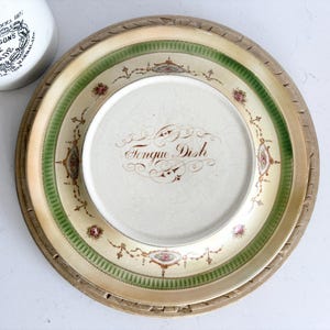 Könnte beinhalten: Eine antike Tongue Dish mit dekorativem Rand, mit einem grün-cremefarbenen Farbschema mit floralen Akzenten. Das Gericht steht auf einem Holztablett, und ein weißes Glas ist teilweise im Hintergrund zu sehen.