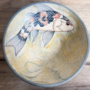 May include: A round ceramic bowl with a grey rim and a speckled yellow and white interior. The bowl features a detailed illustration of a koi fish with black, orange, and white scales swimming across the bottom surface of the vessel.