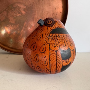 Può includere: Opera d'arte a forma di zucca arancione con dettagli dipinti in nero. La zucca è decorata con un disegno di uccello, con ali e dettagli degli occhi neri. Un vassoio di rame è sullo sfondo.