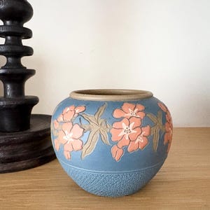 May include: A small, round ceramic vase with a matte blue finish. The exterior features a raised relief pattern of light orange flowers and green leaves encircling the middle. The base has a textured, pebbled surface, and the rim is a neutral tan.