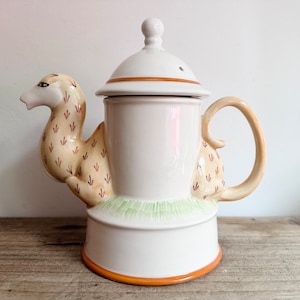 Puede incluir: Una tetera de cerámica blanca con un pico y un asa con forma de camello. La tetera está decorada con manchas marrones y amarillas y tiene una banda verde y blanca alrededor de la base.