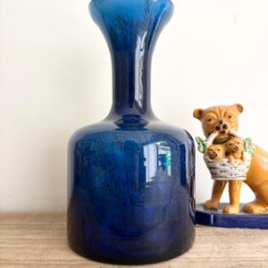 May include: A deep cobalt blue glass vase with a cylindrical body and a flared neck. The glass features subtle dark internal swirls and a smooth, reflective surface. Beside the vase stands a ceramic figurine of a tan dog carrying a basket.