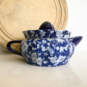 Peut inclure: Une théière en céramique à motifs floraux bleus et blancs avec un couvercle et un bec verseur. La théière a une anse bleu foncé et un couvercle bleu foncé assorti. Une planche à découper en bois est en arrière-plan.