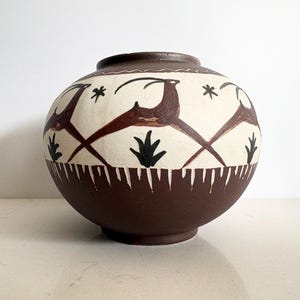 Puede incluir: Un jarrón redondo de cerámica pintado a mano con base y borde marrón. El centro blanquecino presenta diseños de ciervos y estrellas marrones, con formas negras similares a plantas y un borde blanco dentado. El jarrón tiene un acabado mate.