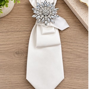 May include: A white satin necktie featuring a large, circular brooch at the center. The brooch is composed of numerous clear, faceted crystals arranged in a starburst pattern, with a cluster of smaller round stones at the very center.