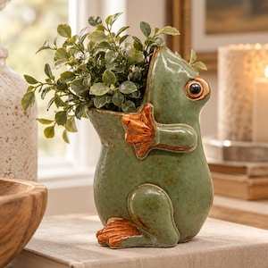 May include: A green ceramic frog-shaped planter with a glossy finish, featuring orange-brown accents on the feet and hands. The frog is depicted in a seated position, holding a small, leafy green artificial plant within its hollow back.