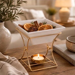 May include: A white square ceramic bowl sits atop a gold-toned metal stand. The bowl contains dried orange slices, cinnamon sticks, and star anise. A small gold-toned metal cup holding a lit white tea light candle is positioned on the base of the stand.