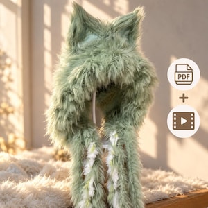 May include: A sage green faux fur hood featuring pointed animal-like ears at the top. The hood has long, dangling side panels with white and green fringe accents. The design includes a PDF icon and a video play icon on the right side of the image.