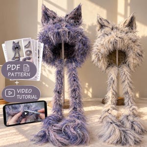 Peut inclure: Deux bonnets à oreilles de chat en fourrure avec de longs rabats, l'un violet et l'autre crème. Inclut un patron PDF imprimé et un tutoriel vidéo sur un écran de smartphone. Les bonnets présentent une texture poilue avec des oreilles pointues.