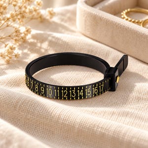 May include: A black plastic ring sizer band featuring gold-colored printed numbers ranging from 5 to 17, with tick marks between each digit. The band includes a small square buckle mechanism with a gold-colored pin to secure the loop.