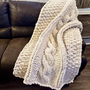 May include: A chunky knit throw blanket in a cream color, featuring a prominent central cable knit pattern flanked by textured moss stitch sections. The blanket is crafted from thick, soft yarn with a visible looped construction.