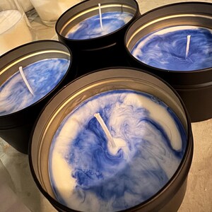 May include: Four round black metal tins containing candles with white and blue marbled wax. Each candle features a centred white cotton wick. The wax surface displays a swirling pattern of deep blue and white colours in a circular motion.