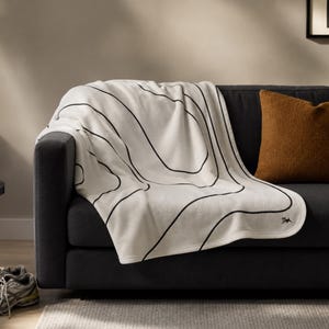 May include: A cream-colored throw blanket featuring a black topographic line pattern draped over a dark gray sofa. The blanket has a small embroidered logo in the bottom corner and is paired with a solid burnt orange square throw pillow.