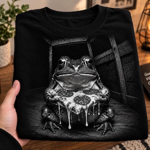 Può includere: Una t-shirt nera con una dettagliata stampa grafica in bianco e nero di una rana seduta su un pavimento di legno, che tiene in mano una fetta di pizza al salamino con formaggio fuso che cola. La rana è posizionata davanti a una struttura in legno.
