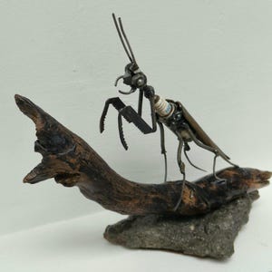 Può includere: Una scultura di mantide religiosa realizzata con componenti metallici, tra cui una candela di accensione per il torace. L'insetto è appollaiato su un pezzo di legno galleggiante scuro e usurato, montato su una base di pietra grigia grezza.