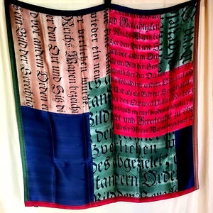 May include: A silk scarf with a patchwork design featuring text in black script. The scarf has sections of pink, green, blue, and beige, with red and green borders. The text appears to be in German.