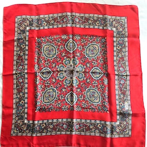 May include: A square scarf featuring a vibrant red border and a central panel with a complex pattern of paisley and floral motifs in shades of blue, yellow, and white. A brown decorative band separates the center from the solid red outer edge.