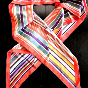 May include: A colorful silk scarf with a vibrant striped pattern. The scarf features a bold red border and a variety of stripes in shades of orange, green, purple, yellow, and white. The scarf is folded and draped, showcasing its design.