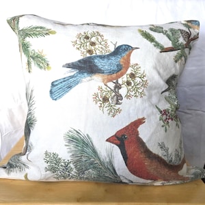 May include: Square decorative pillow with a white background featuring a bluebird and a red cardinal. The birds are perched on branches with green pine needles and small brown pinecones. The pillow is square and appears to be made of fabric.