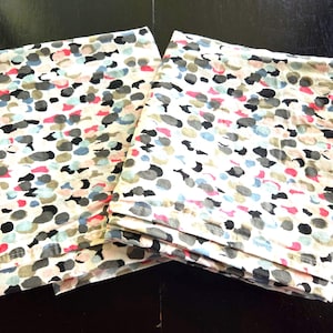 May include: Two folded white fabric items with a colorful pattern of irregular shapes in gray, black, pink, and blue. The items are stacked on a dark surface, showcasing the fabric's design.