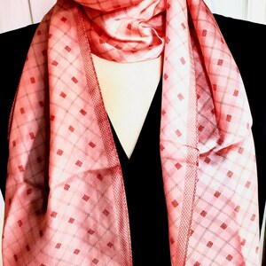 May include: A rectangular scarf in shades of coral and pink featuring a repeating diamond grid pattern with small red square accents. The scarf has a solid red border along the bottom edges and a textured cross-hatch design along the inner vertical panels.