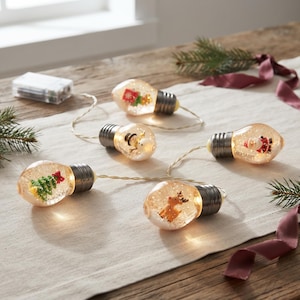Op de afbeelding: Een lichtsnoer met vijf doorzichtige, peervormige ornamenten verbonden door een dunne draad. Elke lamp bevat een miniatuur kerstfiguurtje, waaronder een kerstboom, sneeuwman, rendier, de Kerstman en een cadeautje, omringd door glitters.