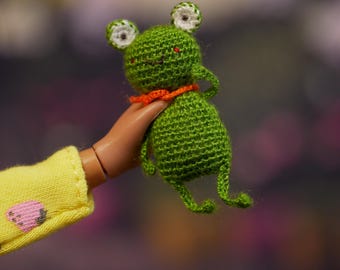 Op de afbeelding: Een handgemaakte groene gehaakte kikkerpop met witte en groene ogen en een oranje kraag. De kikker wordt in een poppenhand gehouden, met een gele mouw zichtbaar. De kikker is ongeveer 7,6 cm hoog.