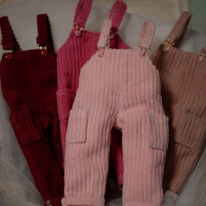May include: Four pairs of corduroy dungarees in different colours: red, pink, light pink, and brown. The dungarees have adjustable straps and pockets.