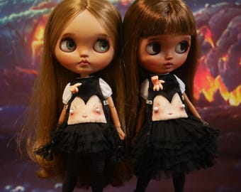 May include: Two Blythe dolls, one with blonde hair and one with brown hair, are dressed in black dresses with a white top and a face design. They wear black boots and stand before a Halloween backdrop.