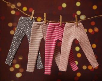 Peut inclure: Quatre paires de leggings pour poupées sont suspendues à une corde à linge. Les leggings présentent différents motifs : gris à pois blancs, rayures roses et grises, rayures roses et rayures rose clair. Ils sont fixés avec des pinces à linge en bois.
