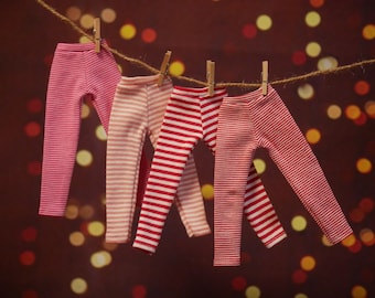 Könnte beinhalten: Vier Paar gestreifte Leggings in Rosa- und Rottönen sind mit Holzklammern an einer Wäscheleine befestigt. Die Leggings hängen vor einem verschwommenen Hintergrund aus warmen, unscharfen Lichtern und schaffen eine gemütliche und einladende Szene.