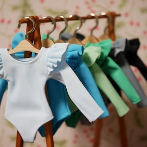 Op de afbeelding: Een set van acht poppen-tops met geruite mouwen die aan een houten rek hangen. De tops zijn in verschillende kleuren, waaronder lichtblauw, turquoise, groen en zwart.