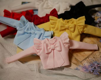 Op de afbeelding: Vijf poppenhemden met geruite kragen en strikken. De shirts zijn in verschillende kleuren, waaronder roze, blauw, geel, rood en wit.
