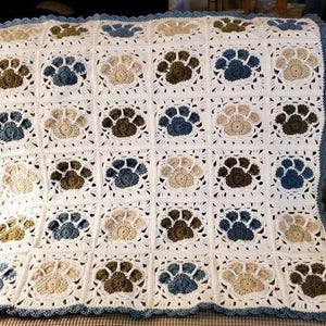 May include: A handmade crochet blanket featuring a repeating grid pattern of white squares, each containing a centered paw print motif in alternating shades of blue, beige, and dark brown. The blanket is finished with a scalloped blue border.