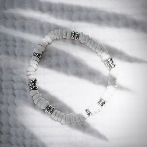 May include: White beaded bracelet with silver-colored metal accents. The bracelet is made of small, irregular-shaped white beads strung together with silver-toned metal spacers. The bracelet is displayed on a white, textured fabric.