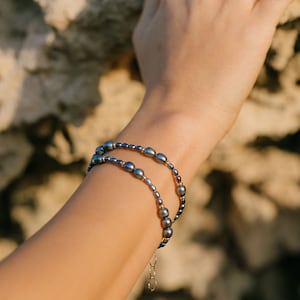 May include: A close-up of a person's wrist wearing two layered bracelets. The bracelets are made of small, dark gray pearls and a thin gold chain. The bracelets are on a light-skinned wrist.