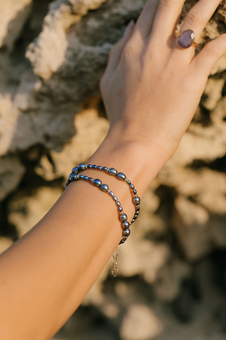 May include: A close-up of a person's wrist wearing two layered bracelets. The bracelets are made of small, dark gray pearls and a thin gold chain. The bracelets are on a light-skinned wrist.