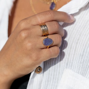 May include: A gold ring with a large, oval-shaped purple gemstone. The ring is worn on a finger with several other gold bands.