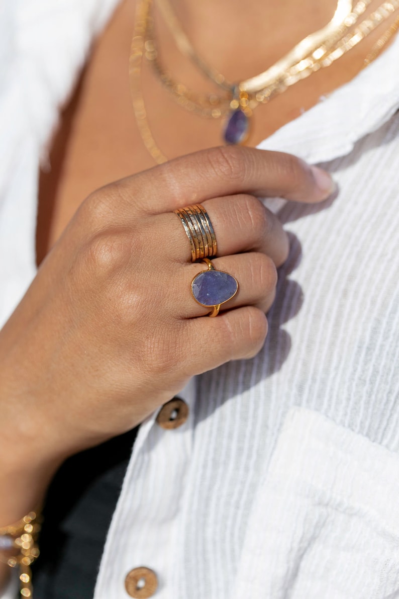 May include: A gold ring with a large, oval-shaped purple gemstone. The ring is worn on a finger with several other gold bands.