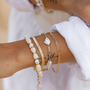May include: A set of four gold bracelets with white pearl accents. The bracelets include a beaded bracelet, a chain bracelet, a delicate chain bracelet with a large pearl, and a simple gold band.