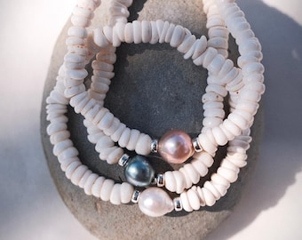 Könnte beinhalten: Drei weiße Muschelbänder mit Perlenakzent. Ein Armband hat eine rosa Perle, eines eine dunkelblaue Perle und eines eine weiße Perle. Die Armbänder liegen auf einem grauen Stein.