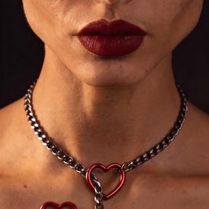 May include: A dark metallic chain necklace featuring a red heart-shaped clasp that connects the chain links. A second red heart-shaped charm is attached to the lower portion of the chain, creating a lariat-style design around the neck area.