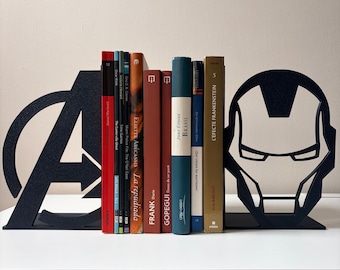 Può includere: Una coppia di fermalibri in metallo nero con il logo degli Avengers a sinistra e la sagoma del casco di Iron Man a destra, che sostengono una fila di vari libri con dorsi di colore rosso, marrone, blu e beige allineati.