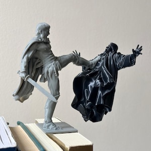 Puede incluir: Una figura impresa en 3D de color gris de un guerrero con capa que sostiene una espada y patea a una figura impresa en 3D de color negro de un personaje con túnica. El guerrero se apoya en una base cuadrada, mientras el otro flota.