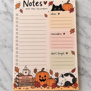 May include: A rectangular notepad featuring an autumn theme with fallen leaves, pumpkins, and cats. The header reads Notes small steps, big progress. It includes a checklist, sections for ideas, reminders, and don't forget, plus a stack of books.