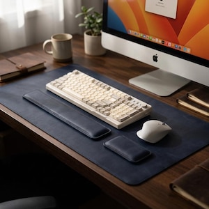 May include: A dark blue leather desk mat featuring a rectangular keyboard, a matching long wrist rest, a smaller rectangular wrist rest, and a light gray computer mouse. The keyboard has light-colored keys and a knob on the top right corner.
