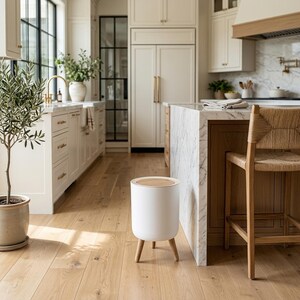 May include: A cylindrical white waste bin with a light wood-toned circular lid, supported by three tapered light wood legs. The bin features a smooth, matte finish and a minimalist design, standing independently on a light-colored wooden floor.