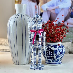 May include: A white ceramic vase with vertical blue stripes, a nutcracker figurine featuring a blue and white pagoda pattern with a bright pink ribbon, and a blue and white porcelain jar filled with artificial red berry branches.